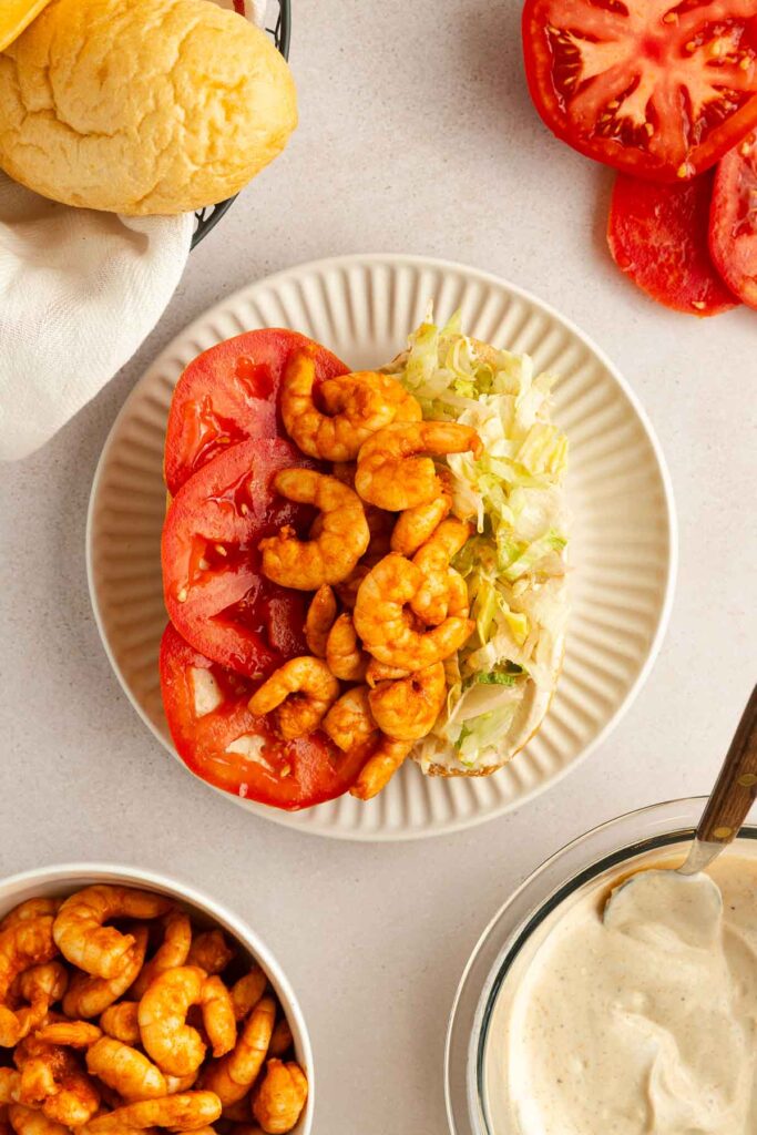 An open-faced shrimp po boy on a plate with sliced tomatoes, shredded lettuce, and seasoned shrimp. Nearby are extra tomato slices, a bowl of shrimp, a bread roll, and a bowl of creamy sauce with a spoon.