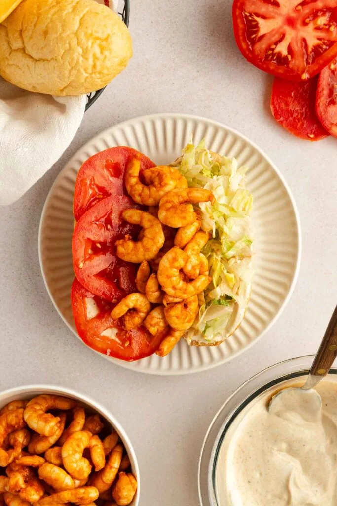 An open-faced shrimp po boy on a plate with sliced tomatoes, shredded lettuce, and seasoned shrimp. Nearby are extra tomato slices, a bowl of shrimp, a bread roll, and a bowl of creamy sauce with a spoon.