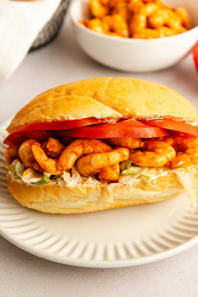 A skinny shrimp po boy sits on a white plate, loaded with shrimp, lettuce, creamy sauce, and sliced tomatoes. A bowl of shrimp is blurred in the background.