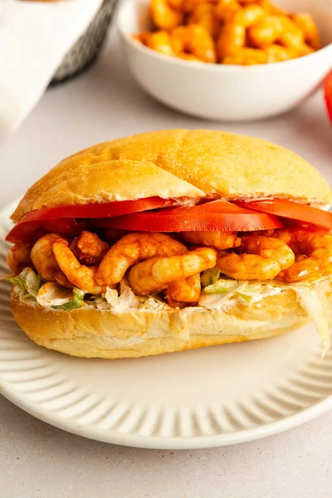 A skinny shrimp po boy sits on a white plate, loaded with shrimp, lettuce, creamy sauce, and sliced tomatoes. A bowl of shrimp is blurred in the background.
