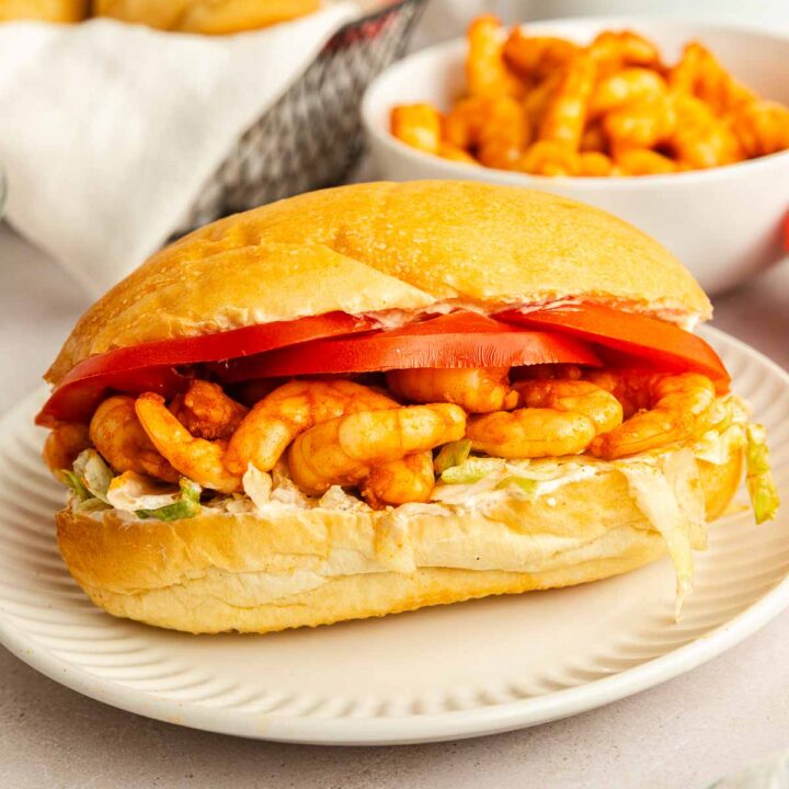 A twist on the classic shrimp po boy, this sandwich is filled with crunchy cheese curls, shredded lettuce, sliced tomatoes, and a creamy spread on a soft bun, served on a white plate with extra cheese curls and buns in the background.