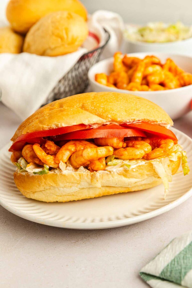 A twist on the classic shrimp po boy, this sandwich is filled with crunchy cheese curls, shredded lettuce, sliced tomatoes, and a creamy spread on a soft bun, served on a white plate with extra cheese curls and buns in the background.