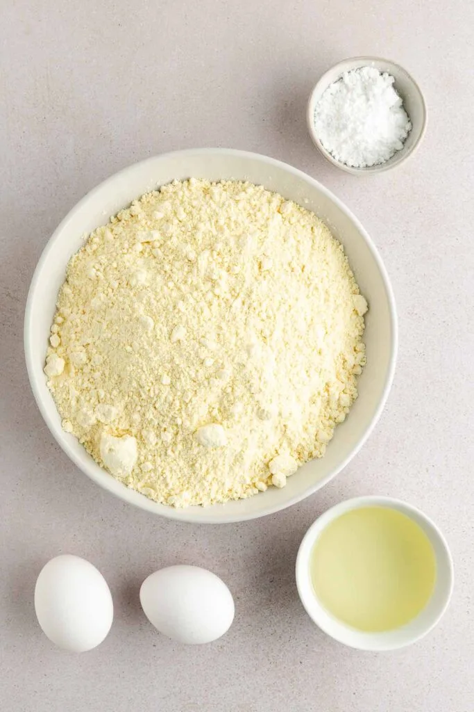 A bowl of yellow cake mix, a small bowl of oil, two eggs, and a small bowl of powdered sugar are arranged on a light countertop.