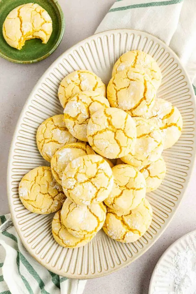 A plate filled with lemon crinkle cookies dusted with powdered sugar sits on a table next to a small green dish holding one cookie, and striped kitchen towels.