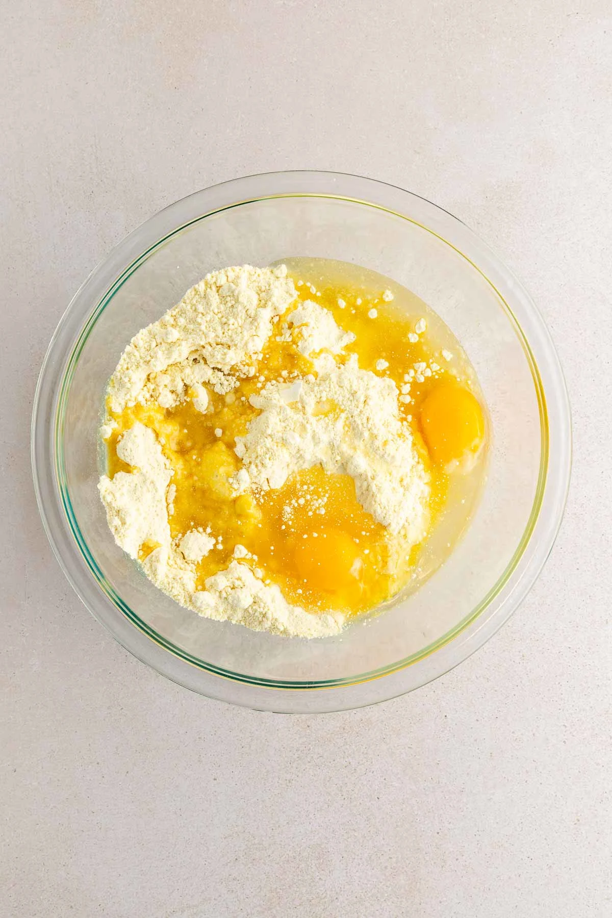 A glass bowl filled with flour, three cracked eggs, and liquid ingredients, all unmixed, sits on a light-colored countertop.