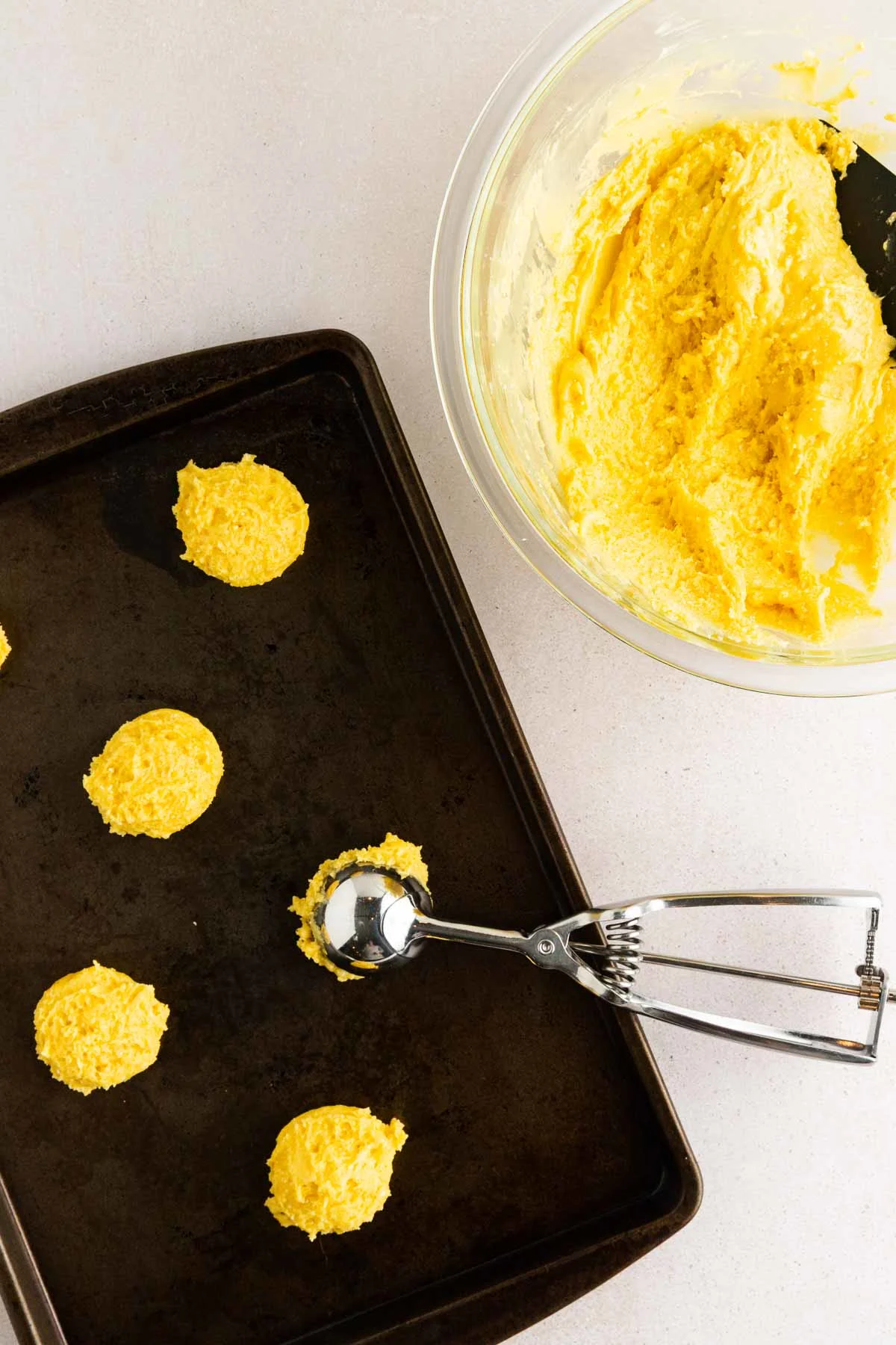A baking sheet with yellow cookie dough scoops, a metal cookie scoop, and a glass bowl filled with more yellow dough on a light surface.