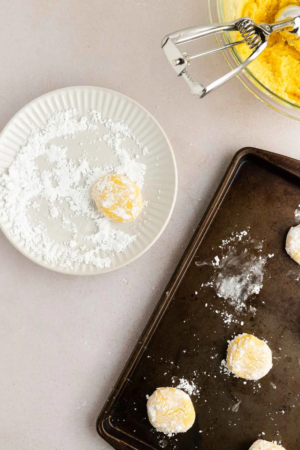 A ball of yellow dough is rolled in powdered sugar on a white plate. A baking tray with sugared dough balls and a glass bowl with dough and a cookie scoop are nearby on a light surface.