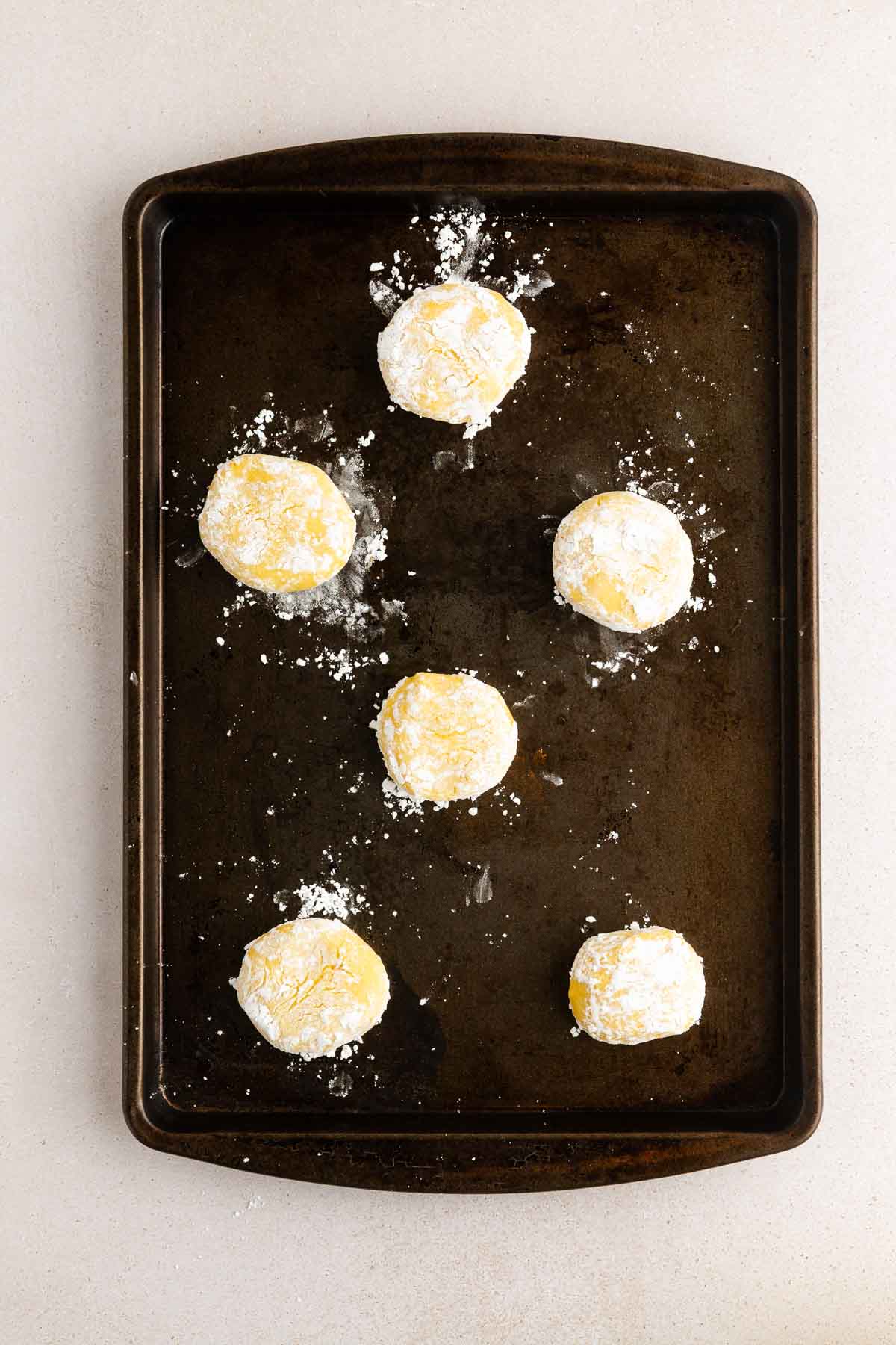 Six unbaked dough balls coated in powdered sugar are arranged on a dark baking sheet, ready to be baked. The baking sheet sits on a light-colored surface.