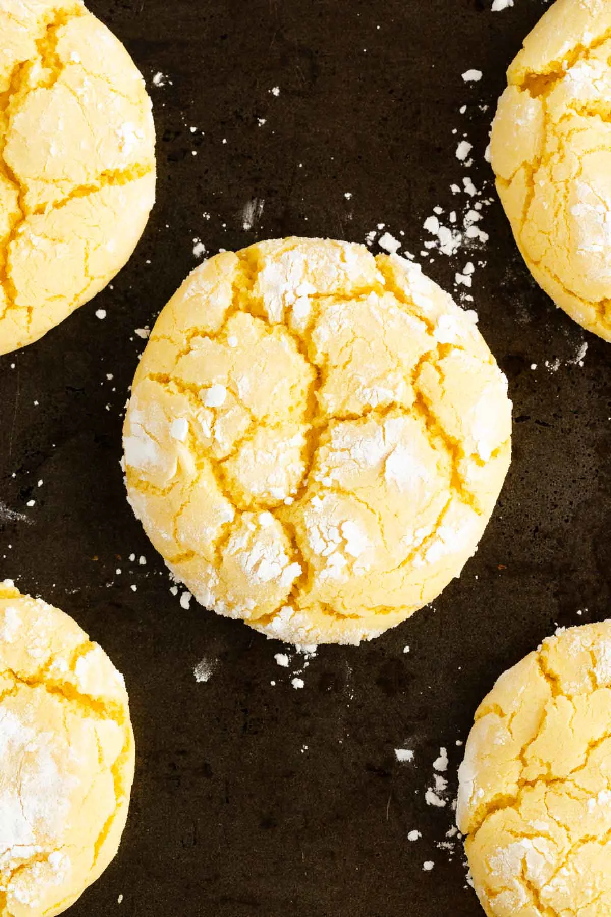 Five lemon crinkle cookies with cracked surfaces and powdered sugar on top, arranged on a dark baking sheet.