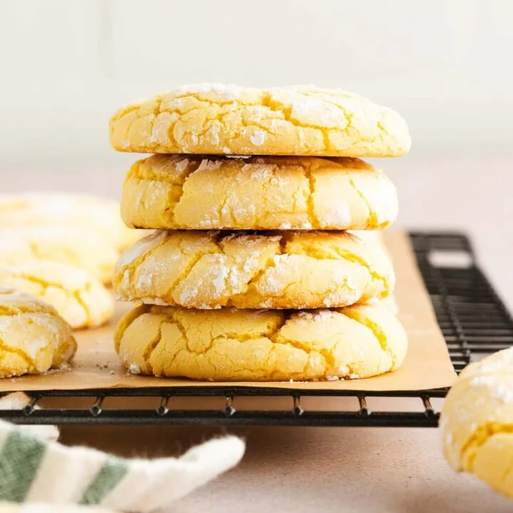 A stack of four lemon crinkle cookies dusted with powdered sugar sits on a wire cooling rack, with more cookies scattered around in the background.