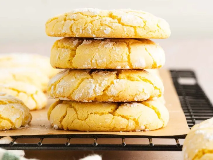 A stack of four lemon crinkle cookies dusted with powdered sugar sits on a wire cooling rack, with more cookies scattered around in the background.