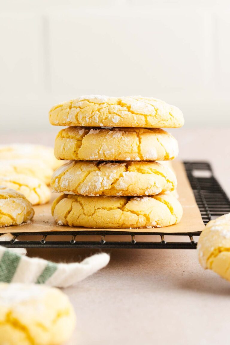 A stack of four lemon crinkle cookies dusted with powdered sugar sits on a wire cooling rack, with more cookies scattered around in the background.