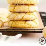 A stack of four powdered sugar-dusted lemon crinkle cookies sits on parchment paper on a cooling rack, with more cookies and a striped cloth partially visible nearby. Text at the bottom reads Lemon Crinkle Cookies.