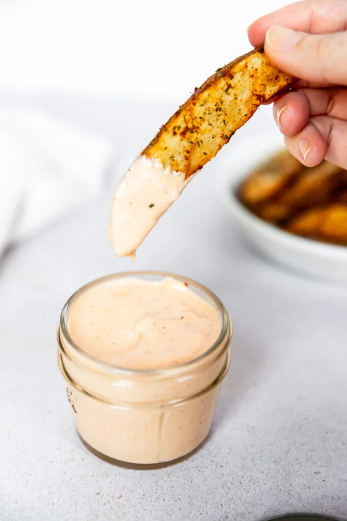 A hand dips a seasoned air fryer potato wedge into a small glass jar of creamy, light orange dipping sauce, with more crispy potato wedges in a bowl in the background.