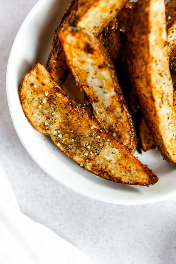 A close-up of seasoned potato wedges air fryer style in a white bowl, garnished with herbs and coarse salt, on a light surface.
