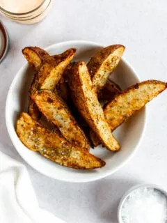 A white bowl filled with seasoned potato wedges in air fryer on a light surface, with a small bowl of salt and a jar of dipping sauce nearby.