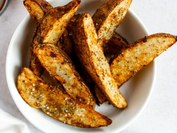 A white bowl filled with seasoned potato wedges in air fryer on a light surface, with a small bowl of salt and a jar of dipping sauce nearby.