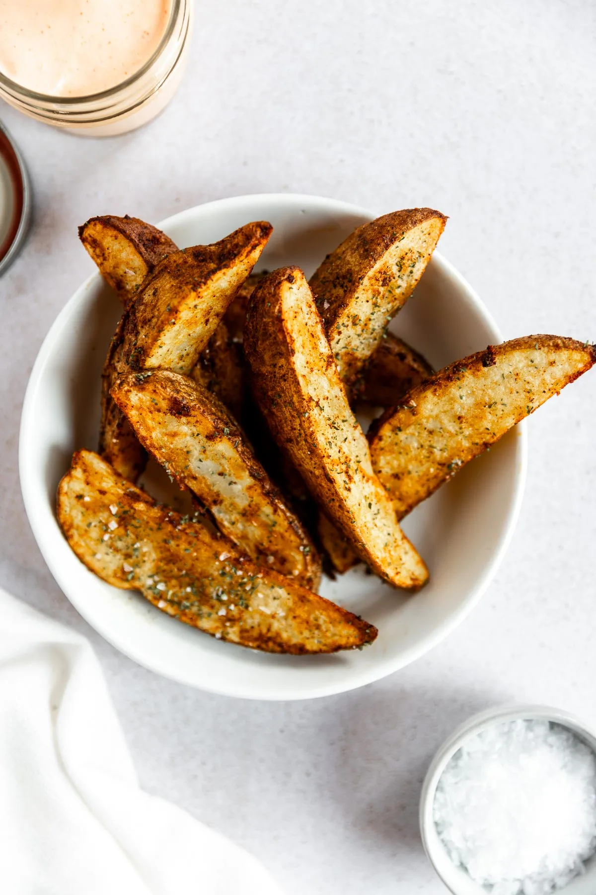 A white bowl filled with seasoned potato wedges in air fryer on a light surface, with a small bowl of salt and a jar of dipping sauce nearby.