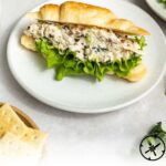 A croissant sandwich filled with chicken salad and lettuce sits on a white plate, with crackers and greens nearby. Text on the image reads: Lemon Basil Chicken Salad - The Culinary Compass.