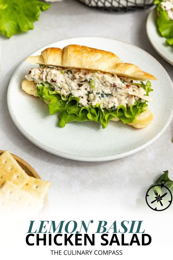 A croissant sandwich filled with chicken salad and lettuce sits on a white plate, with crackers and greens nearby. Text on the image reads: Lemon Basil Chicken Salad - The Culinary Compass.