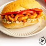 A shrimp po boy sandwich on a white plate, filled with fried shrimp, lettuce, tomato slices, and sauce, with the text Shrimp Po Boy - The Culinary Compass at the bottom.