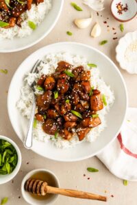 White bowl filled with rice, topped with glossy Air Fryer Honey Garlic Chicken pieces, sliced green onions, and sesame seeds; a fork rests in the bowl. Surrounding the bowl are small dishes with green onions, sesame seeds, garlic, and a honey dipper.