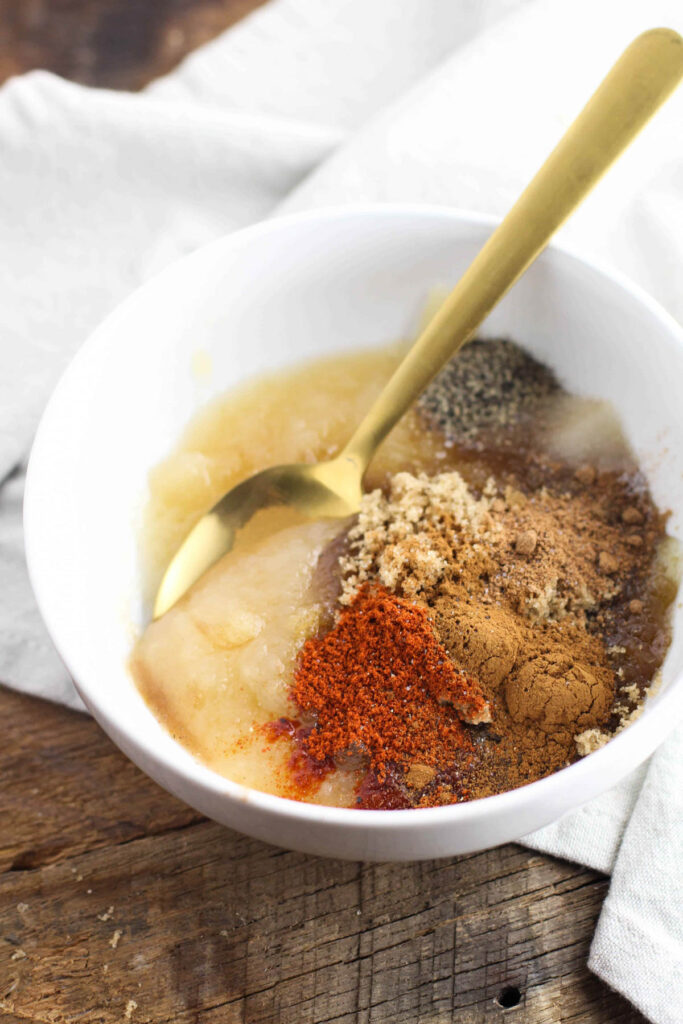 A white bowl contains applesauce, assorted brown and red spices for apple pork chops, and a gold spoon, resting on a wooden surface with a light cloth nearby.