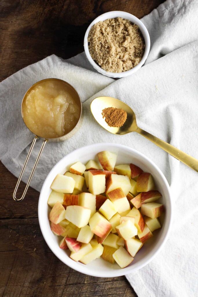 Overhead view of chopped apples in a bowl, a measuring cup of applesauce, a gold spoon with cinnamon, and brown sugarāperfect for preparing delicious apple pork chops on a white cloth and wooden surface.