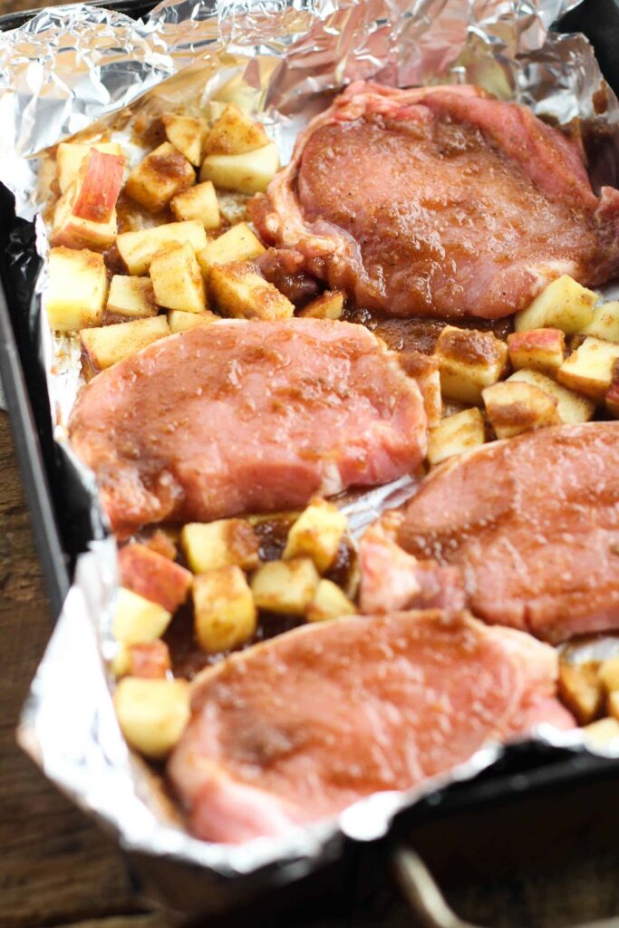 Raw apple pork chops and seasoned diced apples arranged on a foil-lined baking tray, ready to be cooked.