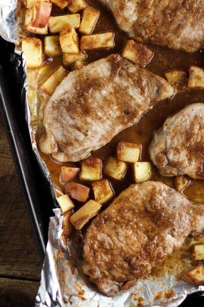 Baked apple pork chops with spices and sauce on a foil-lined tray, surrounded by small diced apple pieces.