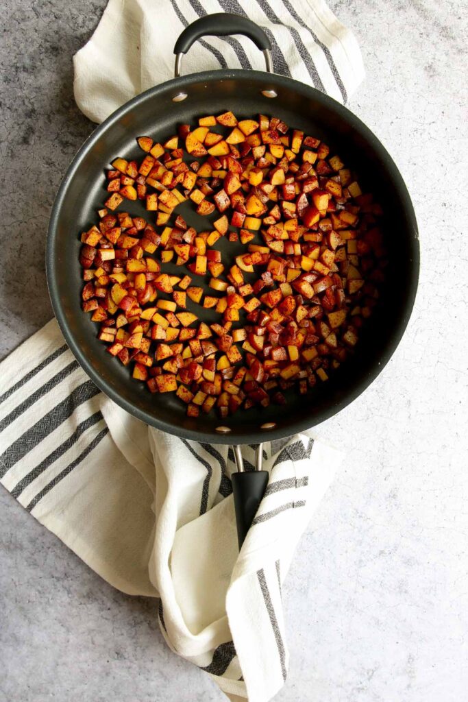 A skillet filled with crispy skillet breakfast potatoes sits on a white and gray striped kitchen towel over a textured gray surface.