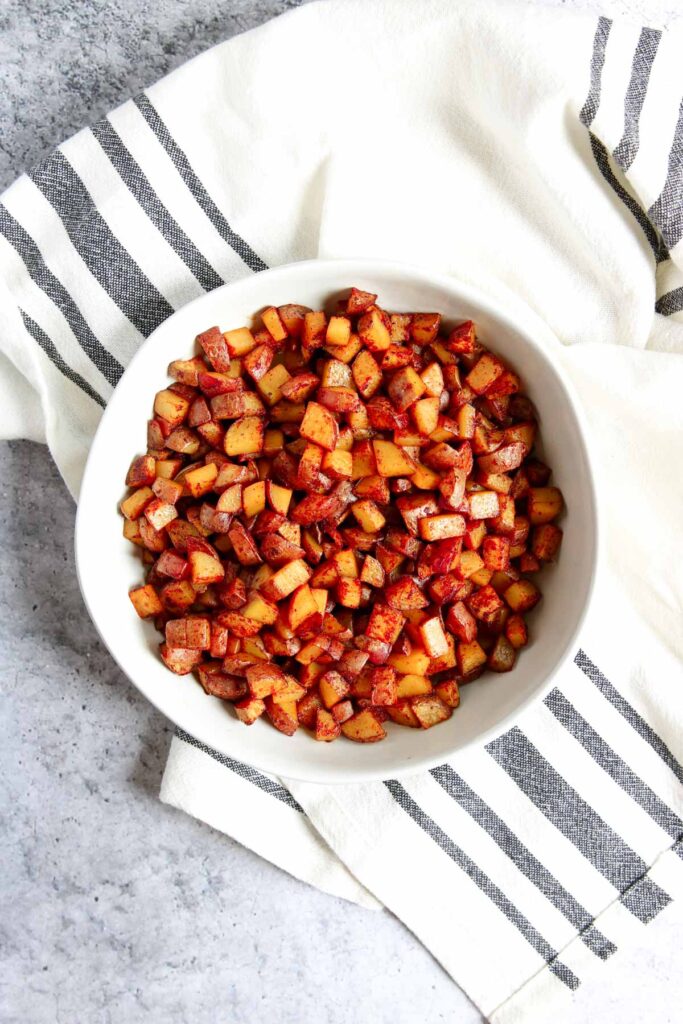 A white bowl filled with crispy skillet breakfast potatoes sits on a white and black striped cloth on a gray surface.