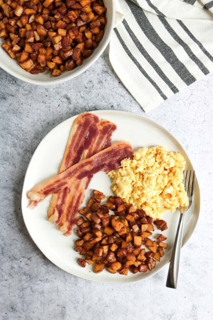 A plate with scrambled eggs, two strips of bacon, and a serving of crispy skillet breakfast potatoes. A fork rests on the plate. A bowl of extra potatoes and a striped towel are nearby on a light surface.