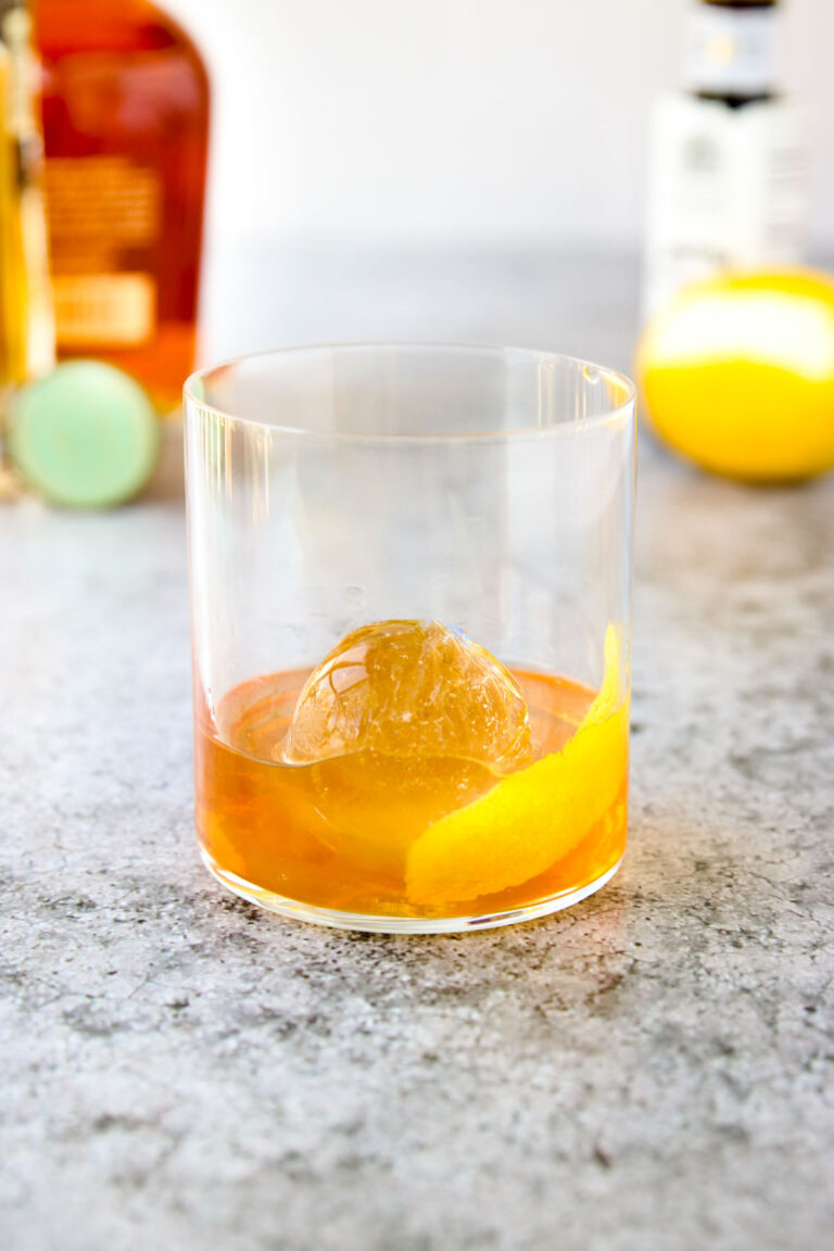 A glass of elderflower old fashioned with a round ice cube and lemon twist garnish sits on a gray surface, with a lemon, a bottle, and bitters blurred in the background.