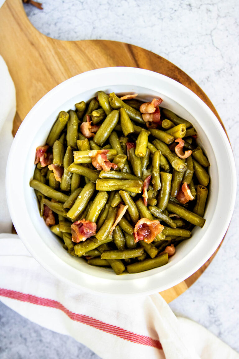 A white bowl filled with cooked green beans and pieces of bacon, reminiscent of a classic green bean bacon casserole, sits on a wooden board with a white and red striped towel nearby.