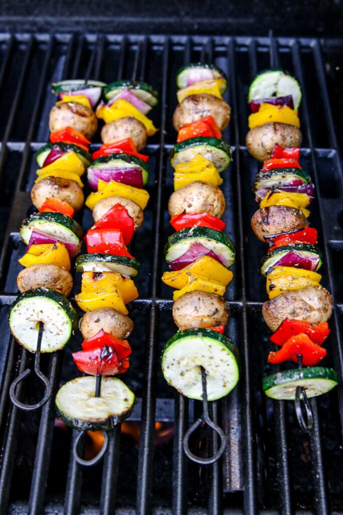 Four grilled veggie skewers with mushrooms, zucchini, red onion, and bell peppers are sizzling on a barbecue. The colorful vegetables are arranged in a repeating pattern on metal skewers, making these grilled veggie skewers as visually appealing as they are delicious.