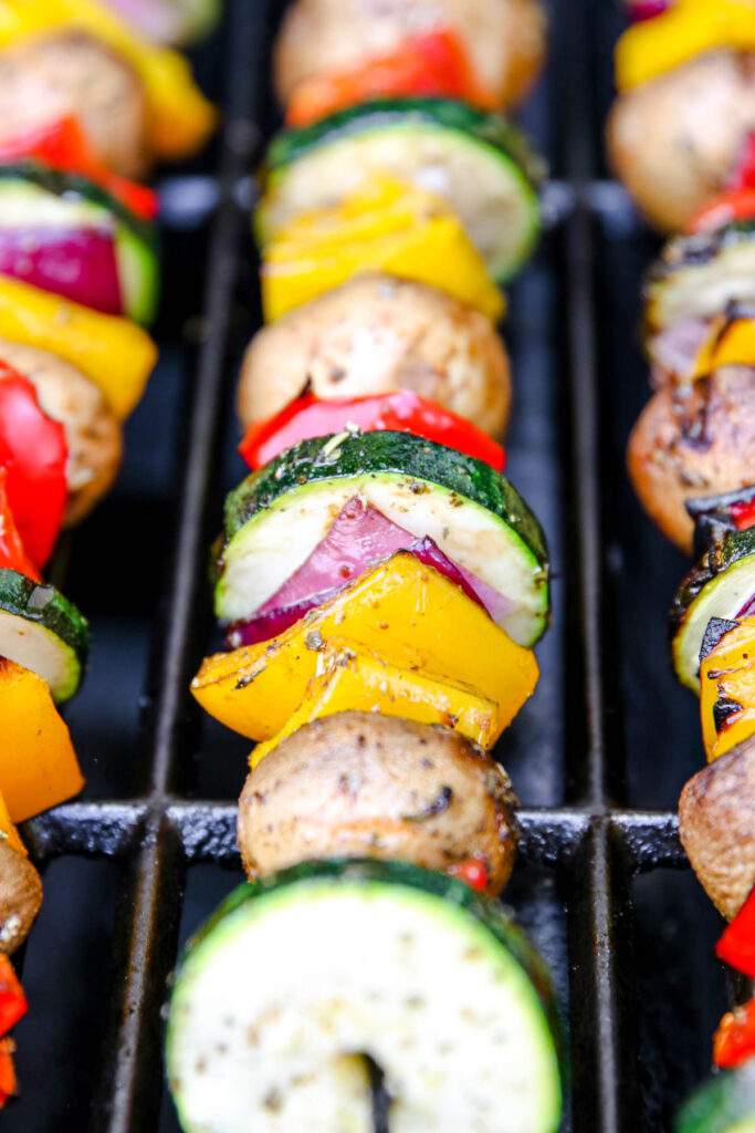 Colorful grilled veggie skewers featuring mushrooms, zucchini, red onion, yellow bell pepper, and red bell pepper sizzle on a barbecue, with perfect grill marks visible on the vegetables.