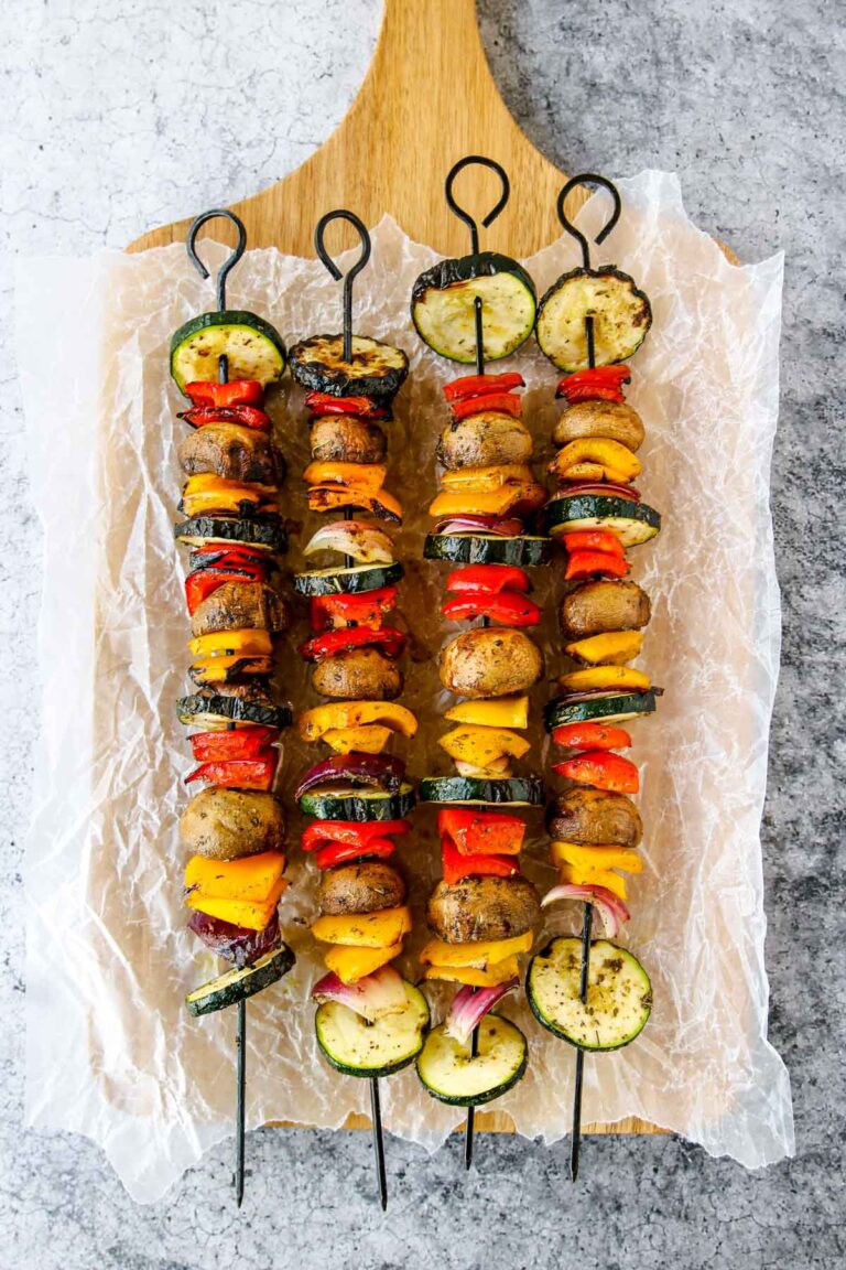 Five grilled veggie skewers with a colorful assortment of zucchini, mushrooms, red onion, bell peppers, and yellow squash are arranged on a wooden board lined with parchment paper.