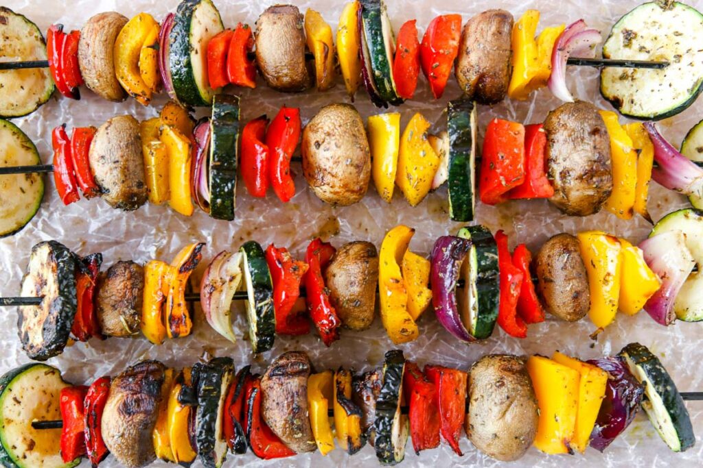 Colorful grilled veggie skewers with mushrooms, zucchini, red onion, and red and yellow bell peppers are artfully arranged on a textured surface, ready to grill or serve. The vegetables appear fresh and perfectly seasoned.