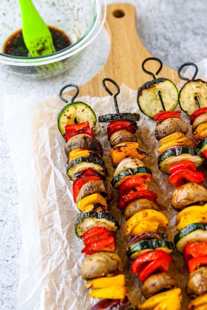 Colorful grilled veggie skewers with zucchini, mushrooms, bell peppers, and onions are arranged on parchment paper over a wooden board. A glass bowl with marinade and a green brush sit nearby.