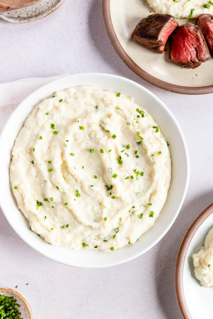 A bowl of creamy horseradish mashed potatoes topped with chopped chives, surrounded by plates of steak and other food on a light surface.