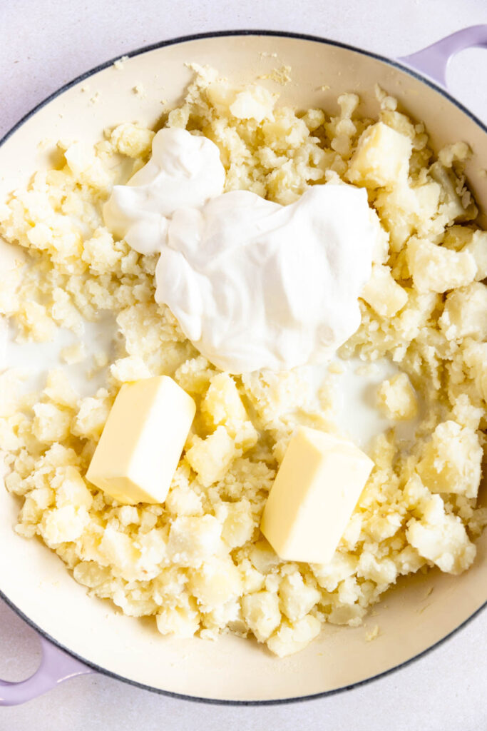 A pot filled with mashed potatoes, two sticks of butter, and a dollop of sour cream, ready to be mixed together into delicious horseradish mashed potatoes.