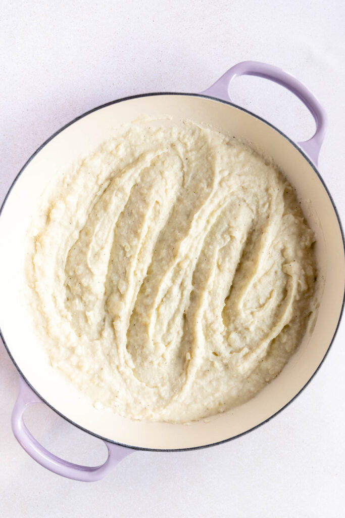 A large pot with light purple handles contains creamy horseradish mashed potatoes, smoothed with visible ridges on top, set on a white surface.