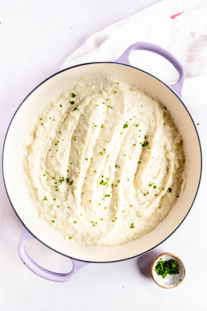 A white pot with light purple handles holds creamy mashed potatoes with horseradish, garnished with chopped green herbs. A small bowl with more herbs sits nearby on a white surface.