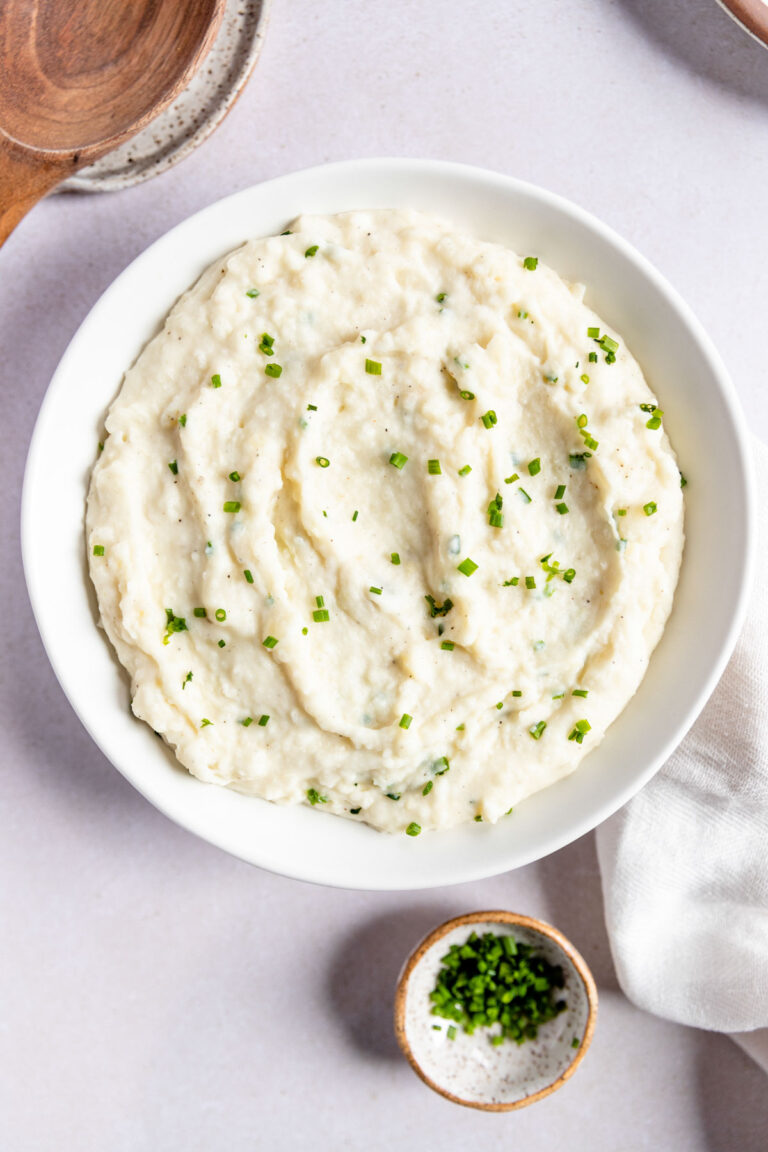 A white bowl filled with creamy mashed potatoes with horseradish, garnished with chopped chives, sits on a light surface next to a small bowl of more chives and a white napkin.