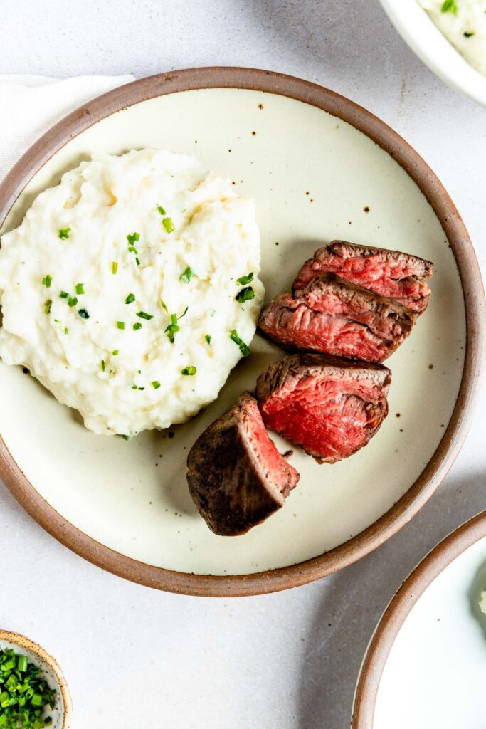A plate with a serving of horseradish mashed potatoes topped with chopped chives and three slices of medium-rare steak, arranged neatly on a light-colored ceramic plate.