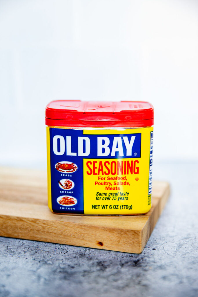 A 6-ounce container of Old Bay Seasoning sits on a wooden board. The label is yellow and blue with red accents, displaying images of crab, shrimp, and chicken—a classic choice when considering Old Bay vs Cajun Seasoning for your next seafood feast.