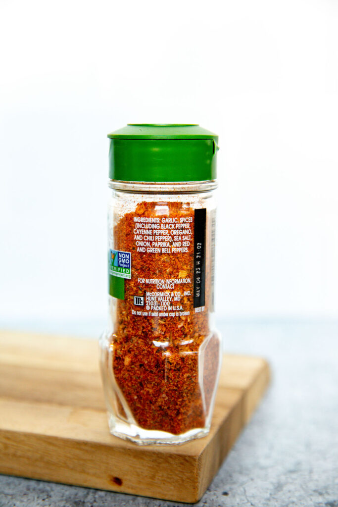 A clear spice jar with a green lid sits on a wooden board. The label shows ingredients and a non-GMO symbol. The jar contains a reddish seasoning mix, perfect for those curious about Old Bay vs Cajun Seasoning. The background is plain and light.