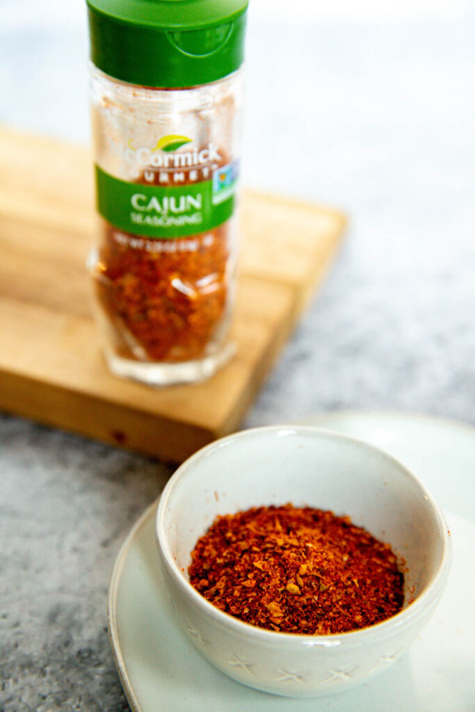 A small bowl of Cajun seasoning sits on a plate, with a bottle of McCormick Cajun Seasoning in the background on a wooden cutting board—perfect for those curious about the flavors and differences when considering Old Bay vs Cajun Seasoning.