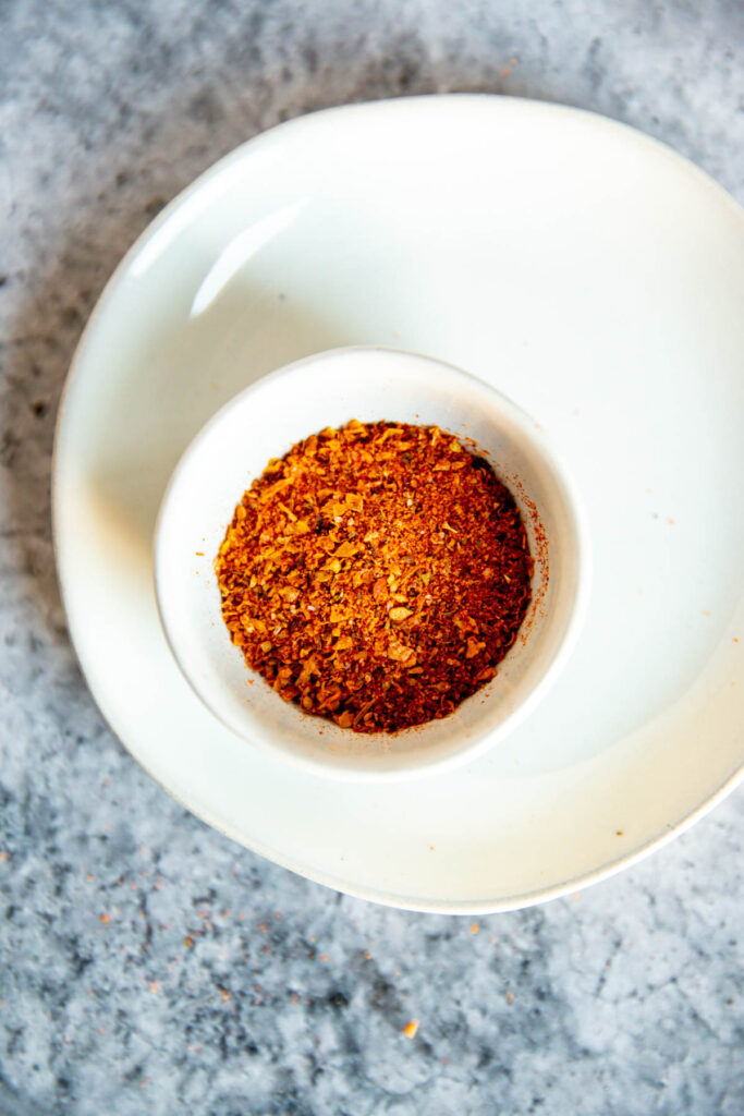 A small white bowl filled with red and orange chili powder sits on a larger white plate, placed on a textured gray surface, evoking the vibrant color contrast often seen when comparing Old Bay vs Cajun Seasoning.